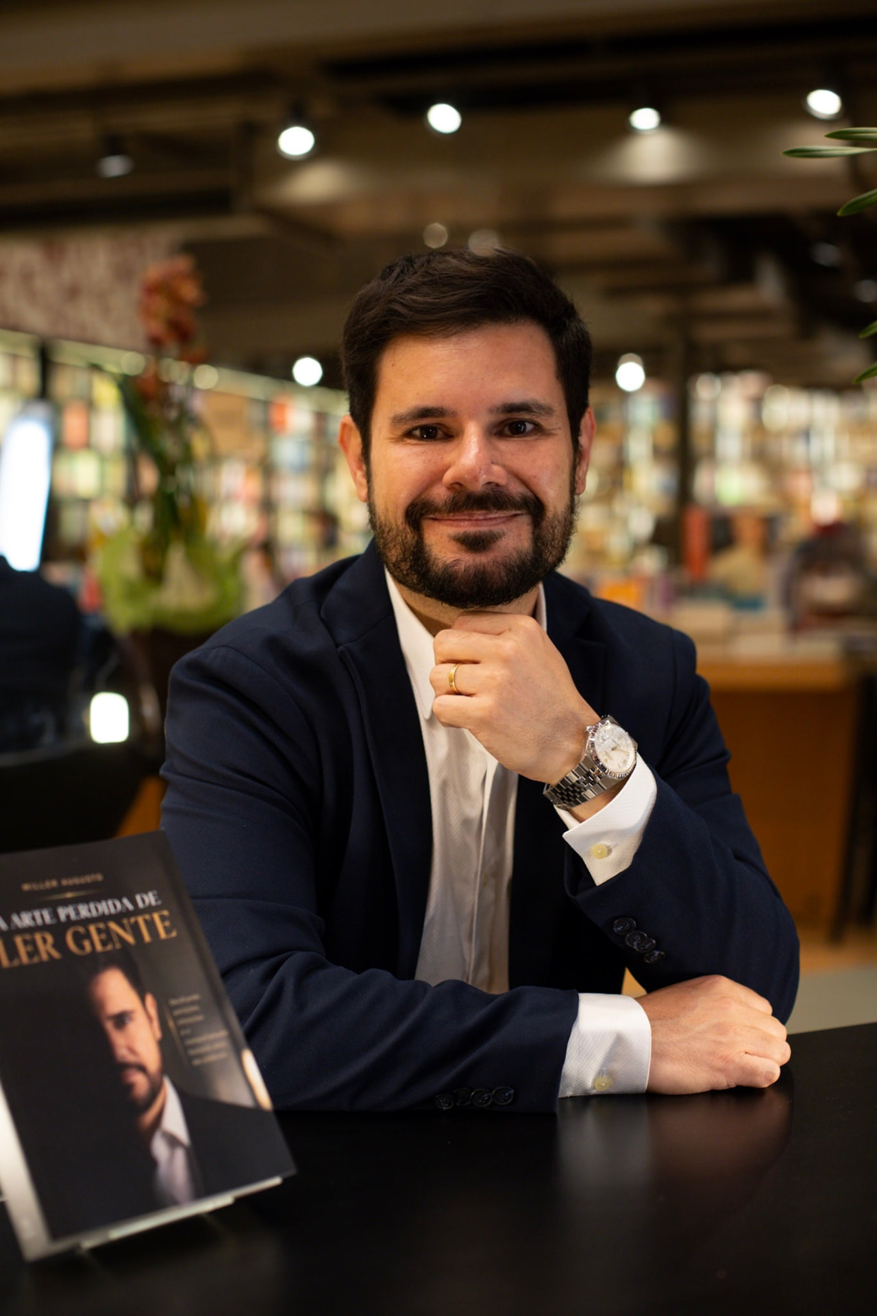 Miller Augusto sentado em livraria ao lado de seu livro